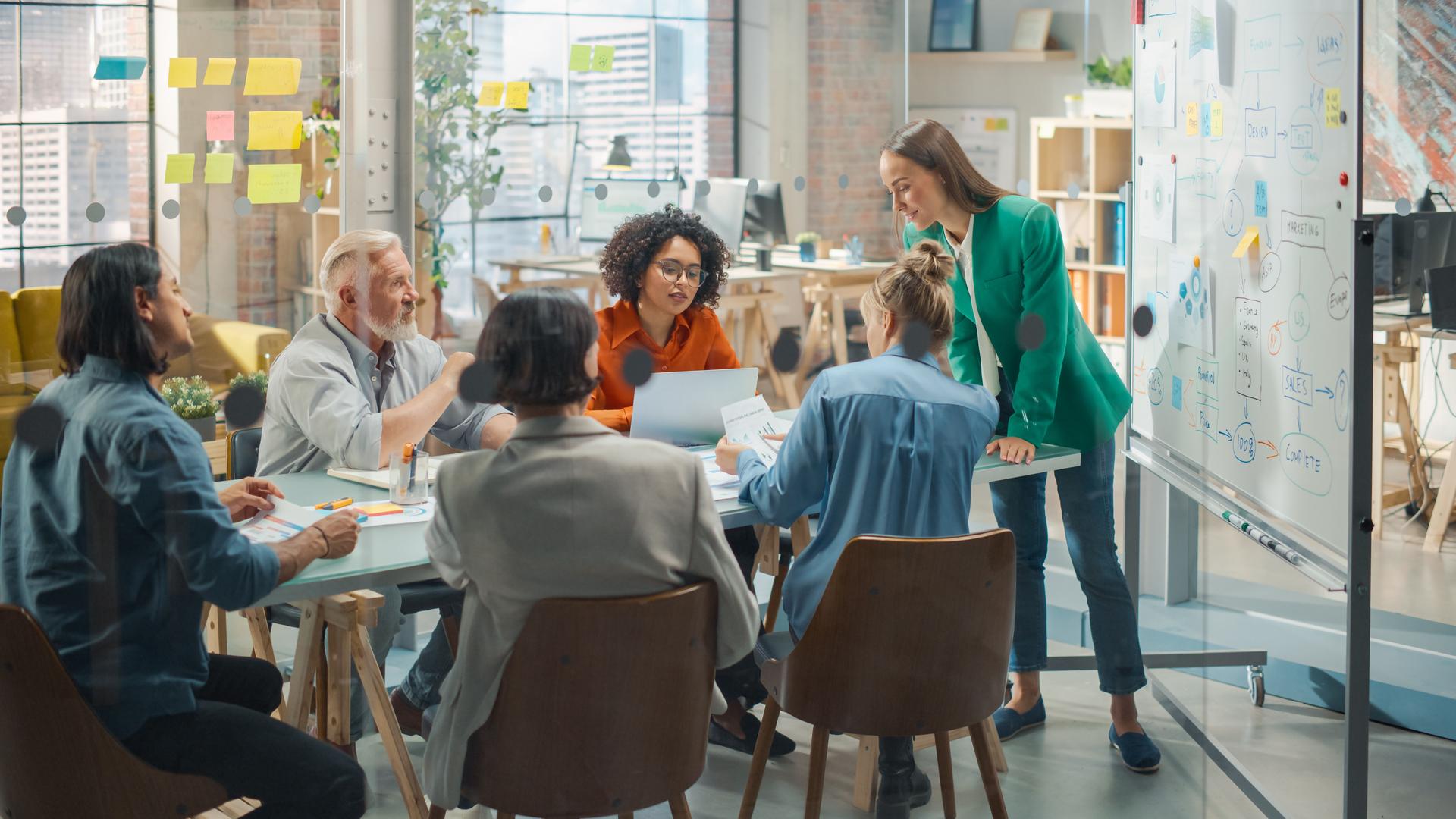 Female Chief Analyst Holds Meeting Presentation for a Team of Economists. She Shows a Whiteboard with Growth Analysis, Charts, Statistics and Data, Answers Questions. People Work in Creative Office